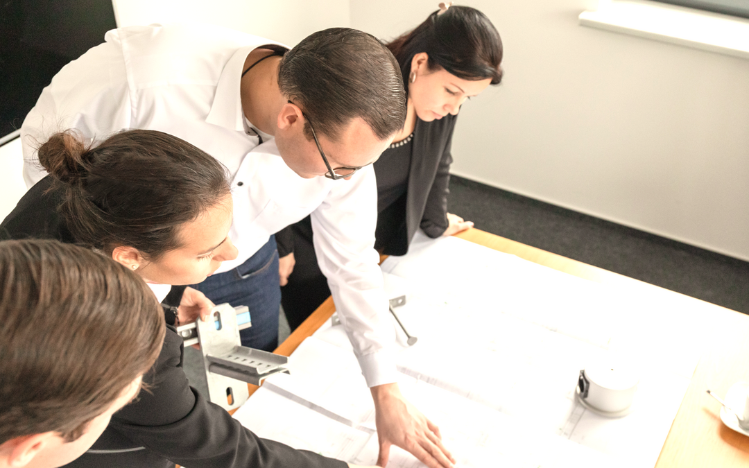 Vier Personen beugen sich über einen Tisch und deuten auf dort ausgebreitete Dokumente.