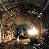 Dunkler, nasser Tunnel mit rauen Wänden, Bauarbeiten und Baufahrzeug, mit Lichtquelle rechts und Wasserpfützen auf dem Boden