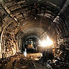 Dunkler, nasser Tunnel mit rauen Wänden, Bauarbeiten und Baufahrzeug, mit Lichtquelle rechts und Wasserpfützen auf dem Boden