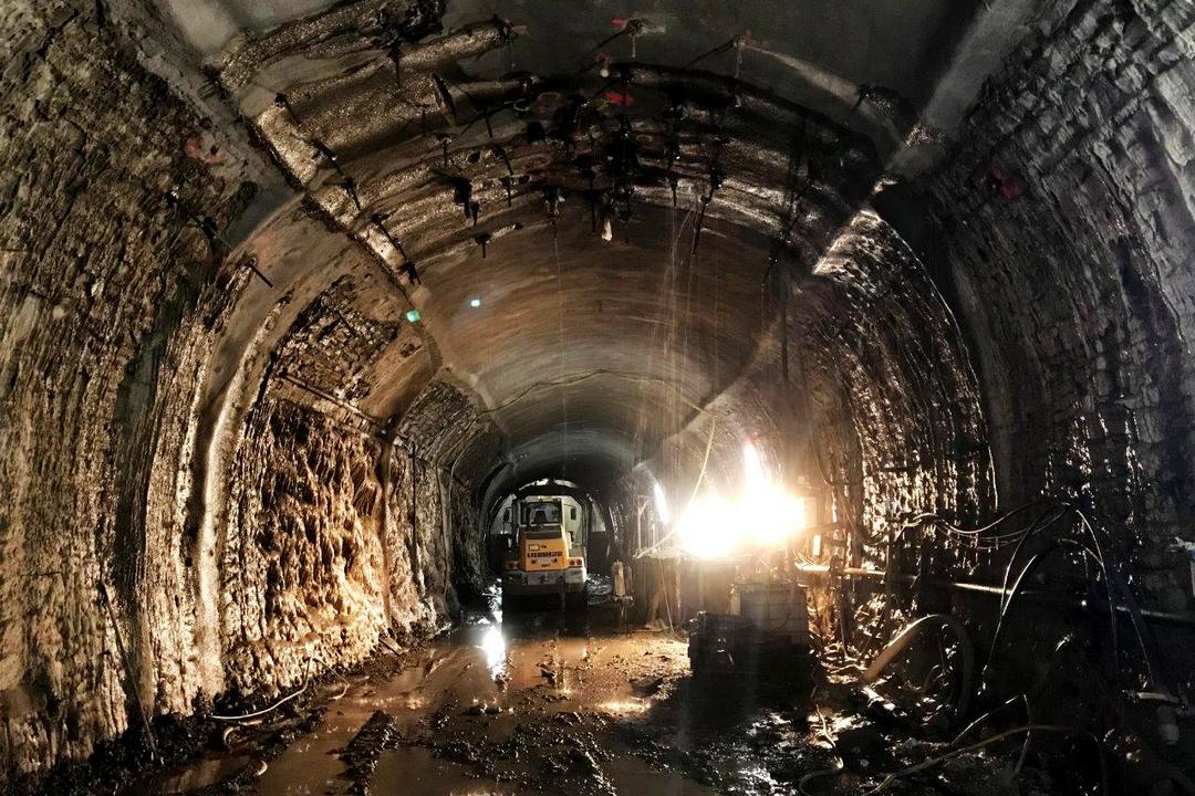 Dunkler, nasser Tunnel mit rauen Wänden, Bauarbeiten und Baufahrzeug, mit Lichtquelle rechts und Wasserpfützen auf dem Boden