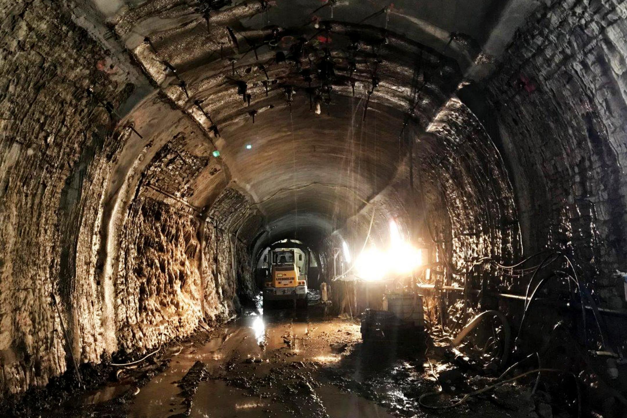 Dunkler, nasser Tunnel mit rauen Wänden, Bauarbeiten und Baufahrzeug, mit Lichtquelle rechts und Wasserpfützen auf dem Boden