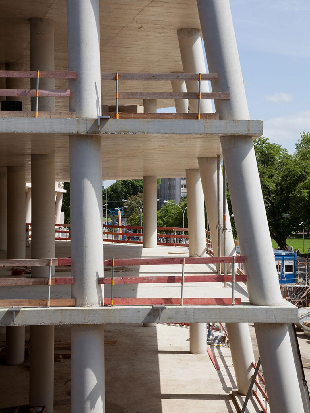 Nahaufnahme eines Rohbaus mit schrägen und geraden Betonstützen sowie provisorischen Holzgeländern
