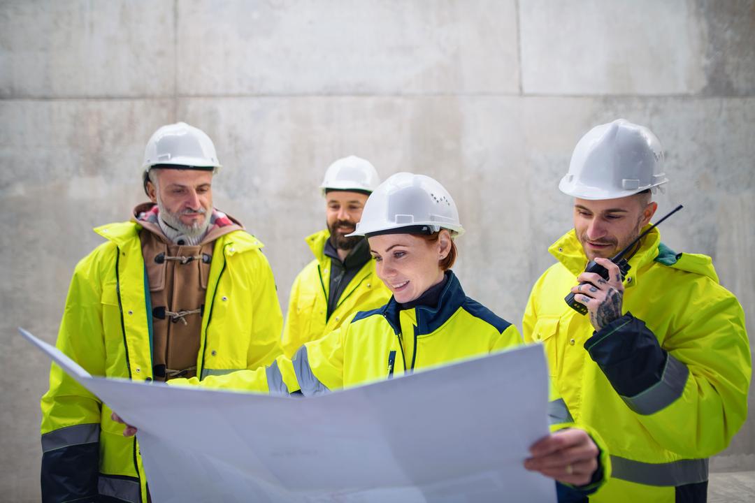 Gruppe von Arbeitern mit Helmen und Warnwesten sieht sich auf einer Baustelle einen Plan an.