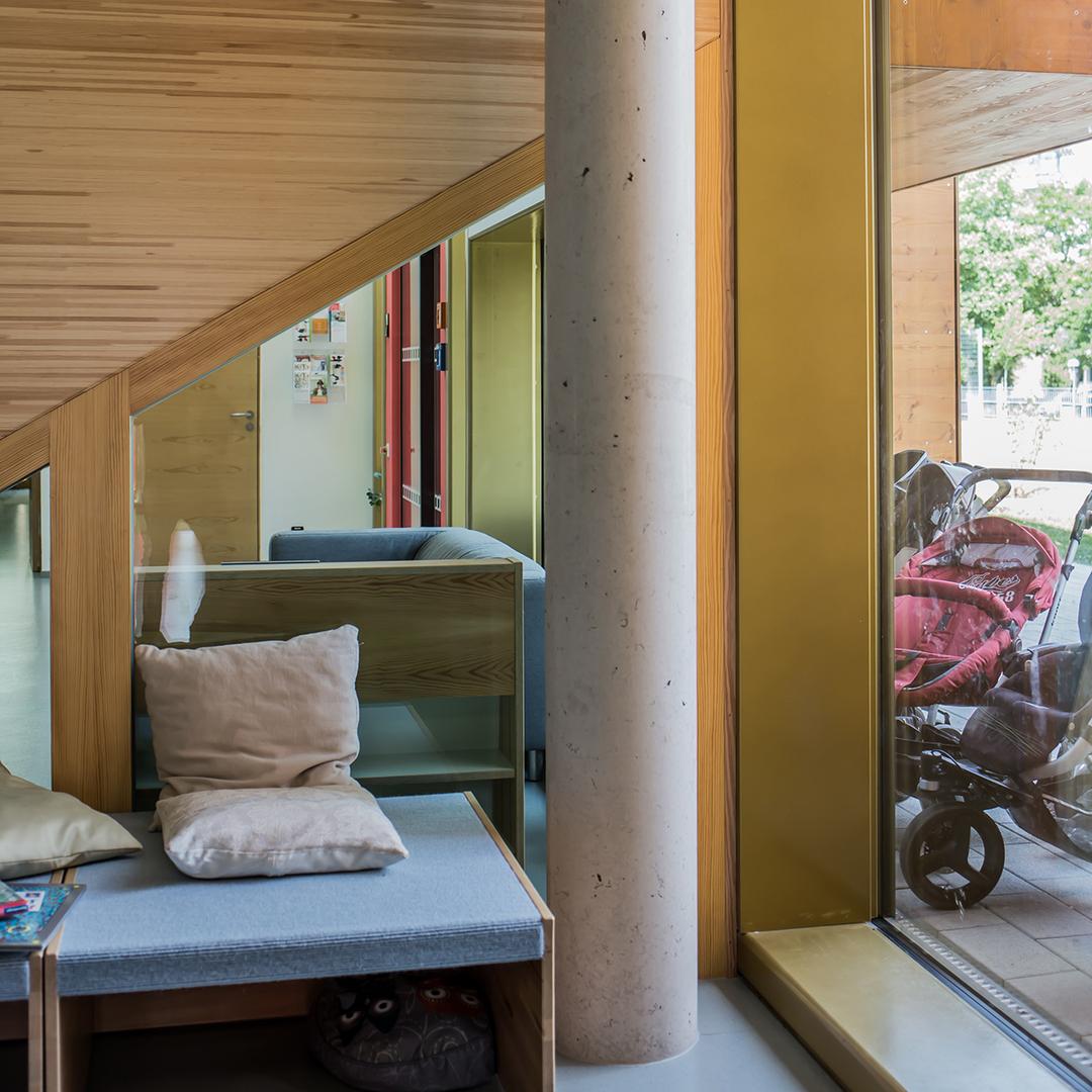 Innenraum mit Holzbank, Betonpfeiler, Glasfront und Blick auf draußen geparkte Kinderwagen