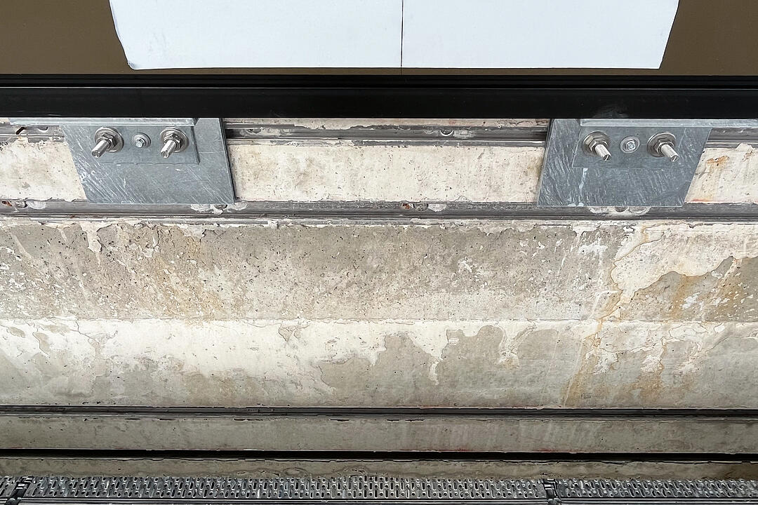 Metallhalterung mit Schrauben an Betonwand, darüber braunes Bauelement mit Klebestreifen.