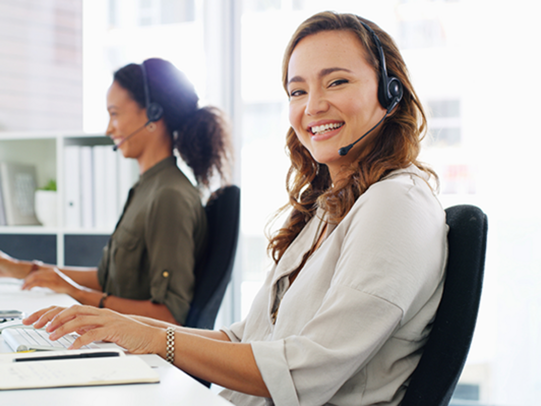 Zwei Frauen mit Headsets sitzen an einem Schreibtisch, eine von ihnen lacht in die Kamera