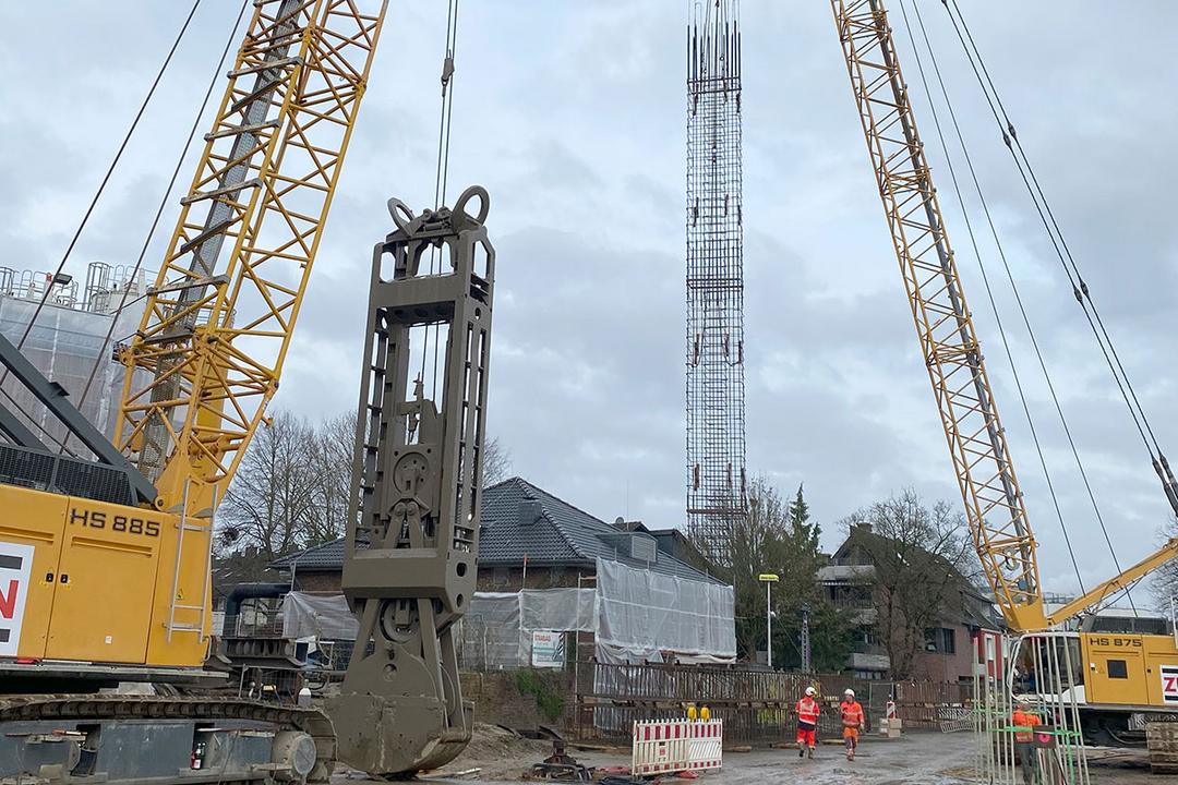 Ein Kran hebt einen hohen Bewehrungskorb auf einer Baustelle, während zwei Bauarbeiter in Sicherheitskleidung vorbeigehen