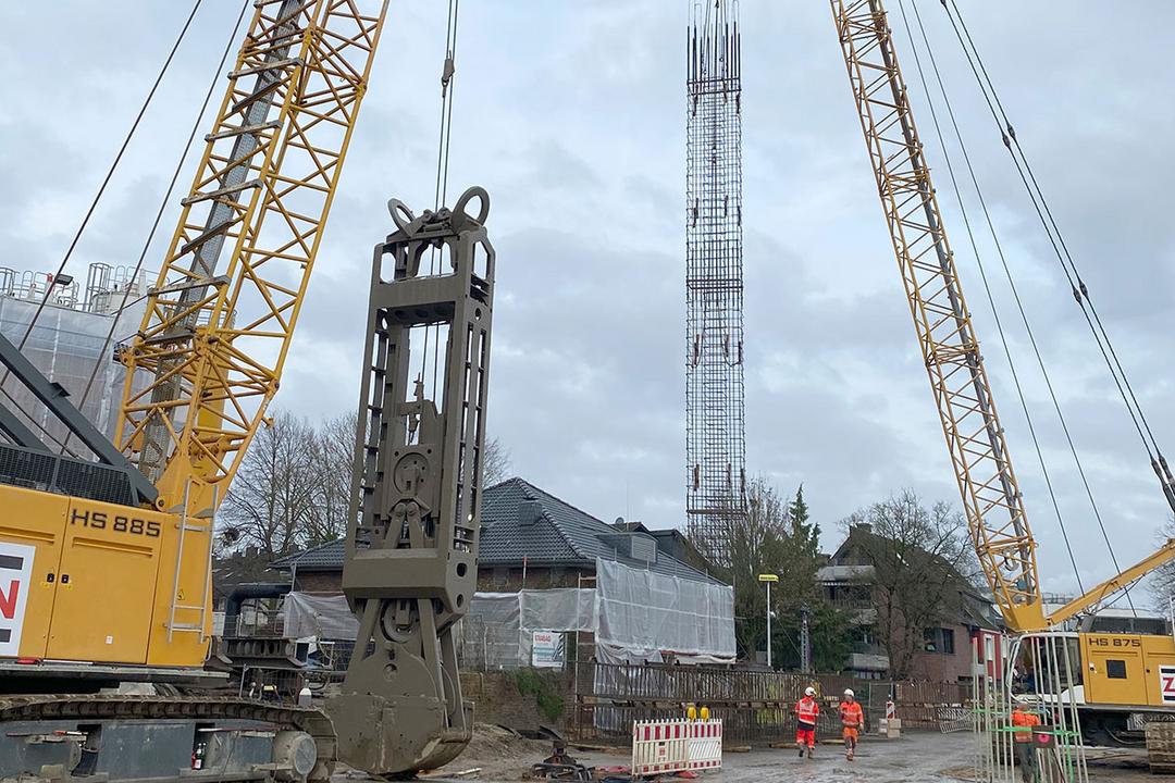 Ein Kran hebt einen hohen Bewehrungskorb auf einer Baustelle, während zwei Bauarbeiter in Sicherheitskleidung vorbeigehen