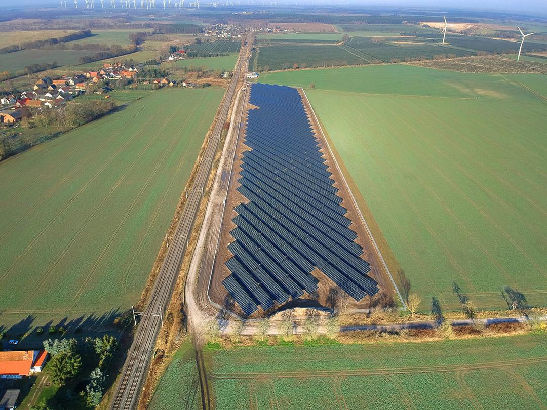 Luftaufnahme eines Solarparks mit Windrädern im Hintergrund, umgeben von Feldern, Bahnstrecke und Dorf