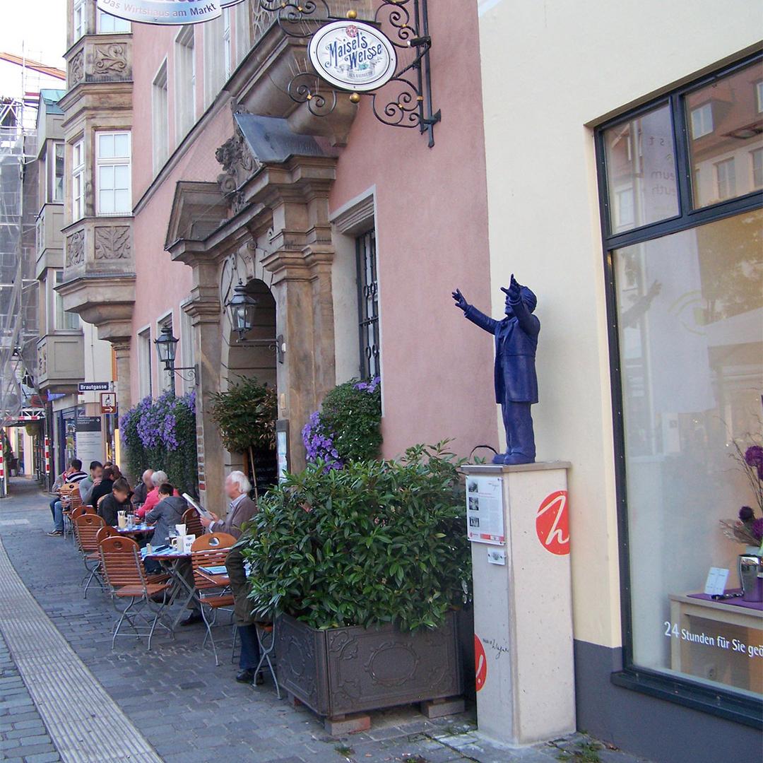 Straßenszene mit Café-Gästen vor historischem Gebäude und blauer Statue eines Dirigenten auf Sockel.