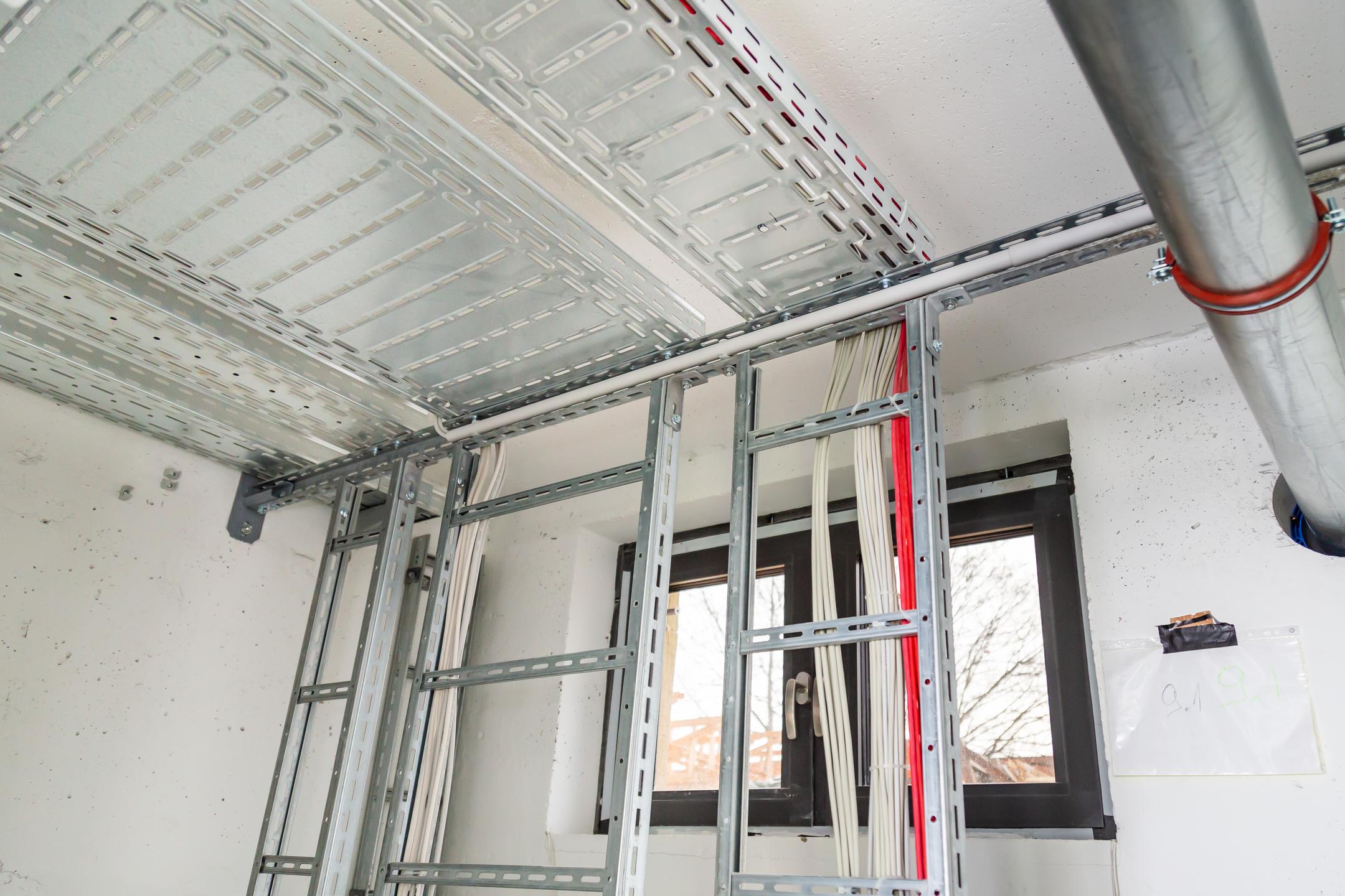 Installierte Kabeltrassen mit geführten Leitungen an Wand und Decke in einem Technikraum mit Fenster und sichtbaren Rohrleitungen