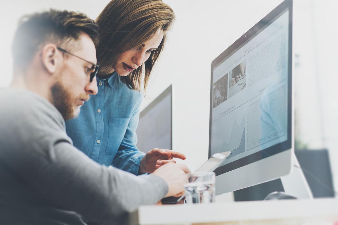 Zwei Arbeitskollegen, eine Frau und ein Mann, sehen sich an einem Schreibtisch etwas zusammen auf einem Computer-Monitor an