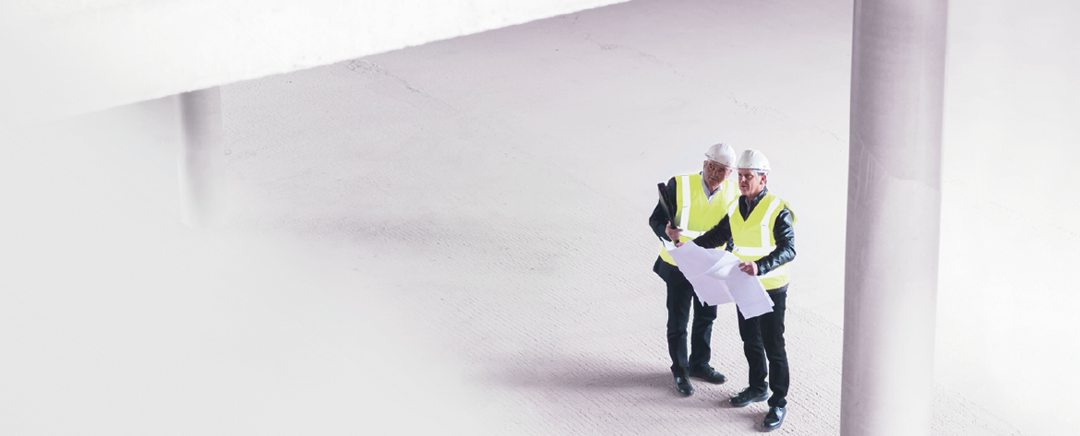 Zwei Männer in gelben Westen halten einen Plan und schauen nach oben.