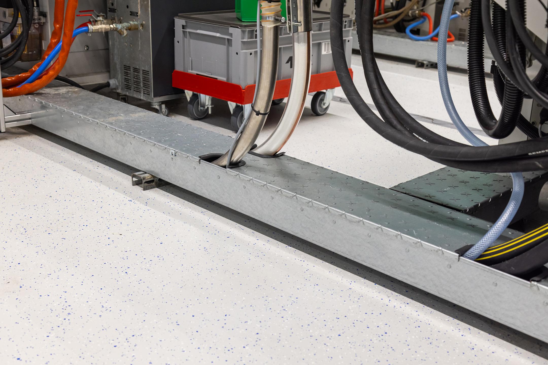 Detailansicht begehbarer Bodenkanal mit Produktionsmaschinen im Hintergrund.