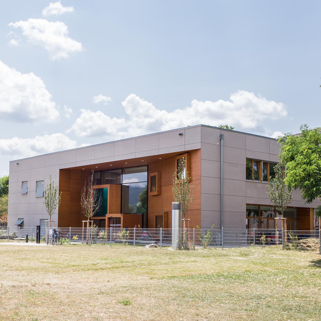 Modernes zweistöckiges Gebäude mit großen Fenstern, umzäunt und von Bäumen umgeben, unter Wolkenhimmel
