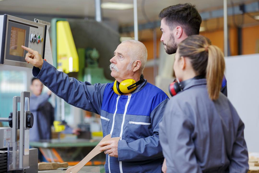 Ein älterer Produktionsmitarbeiter zeigt zwei jüngeren Mitarbeitern etwas an einer Maschine