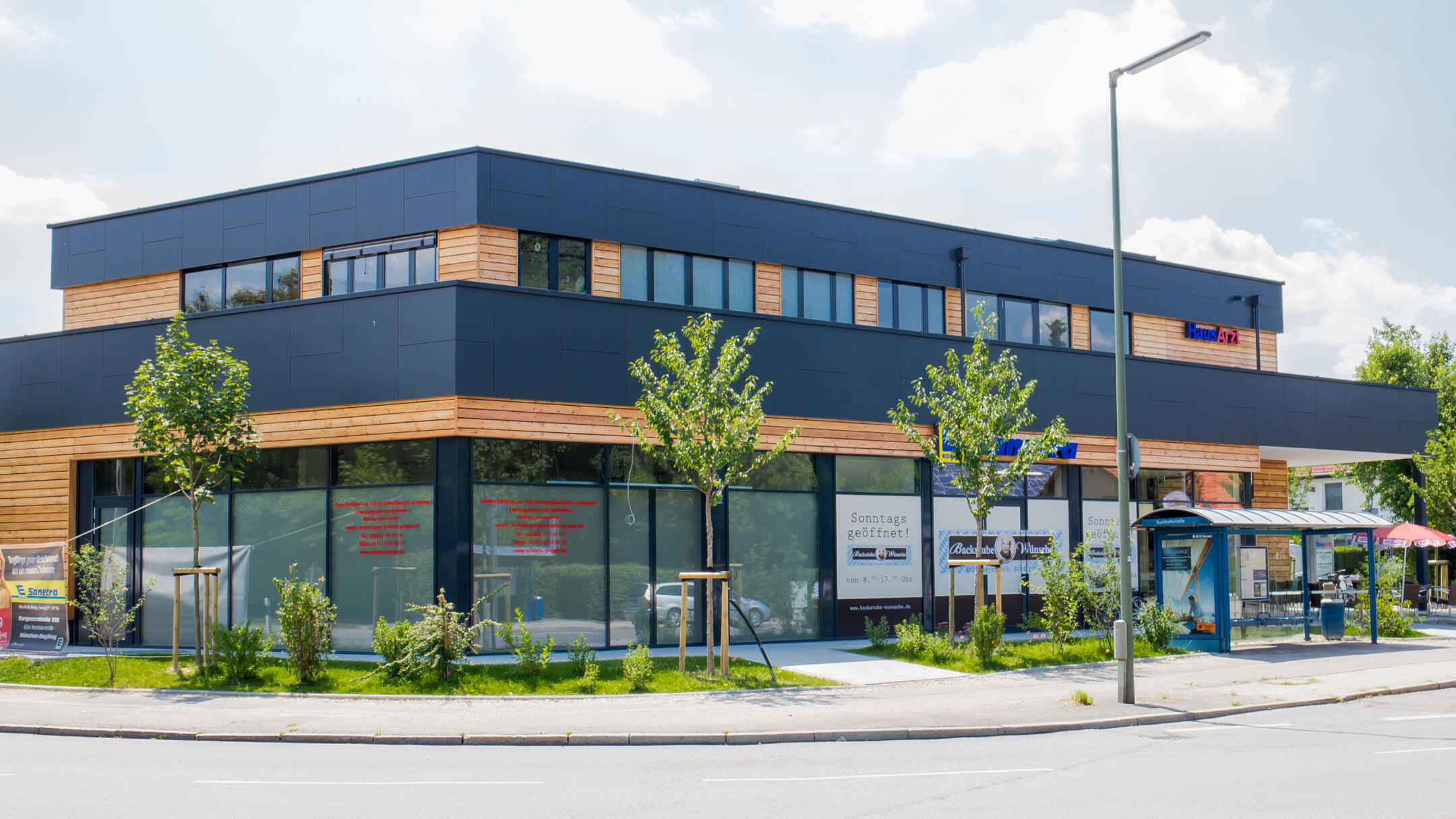 Modernes Gebäude mit großen Fenstern und Bushaltestelle davor, Firmenschilder sichtbar, leicht bewölkter Himmel