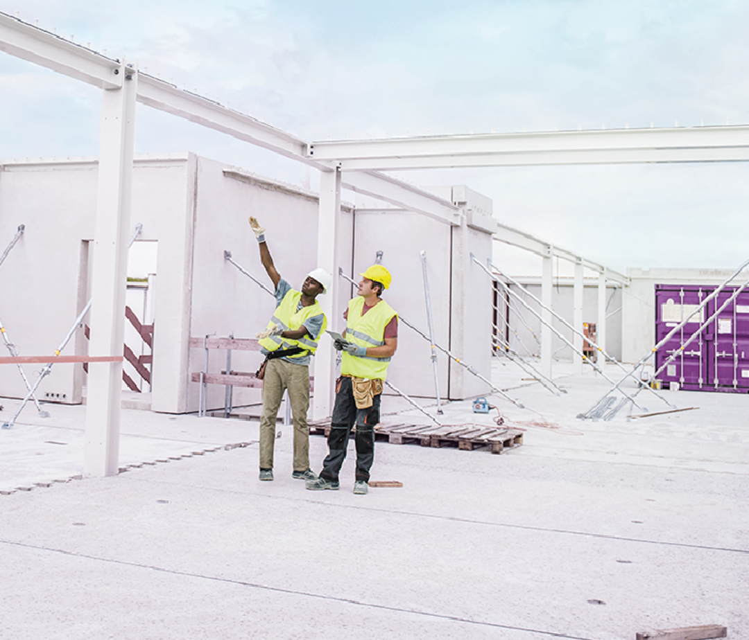 Zwei Personen mit gelben Westen und Helmen auf dem Kopf stehen auf einer Baustelle. Einer zeigt auf etwas, der andere schaut hinterher.