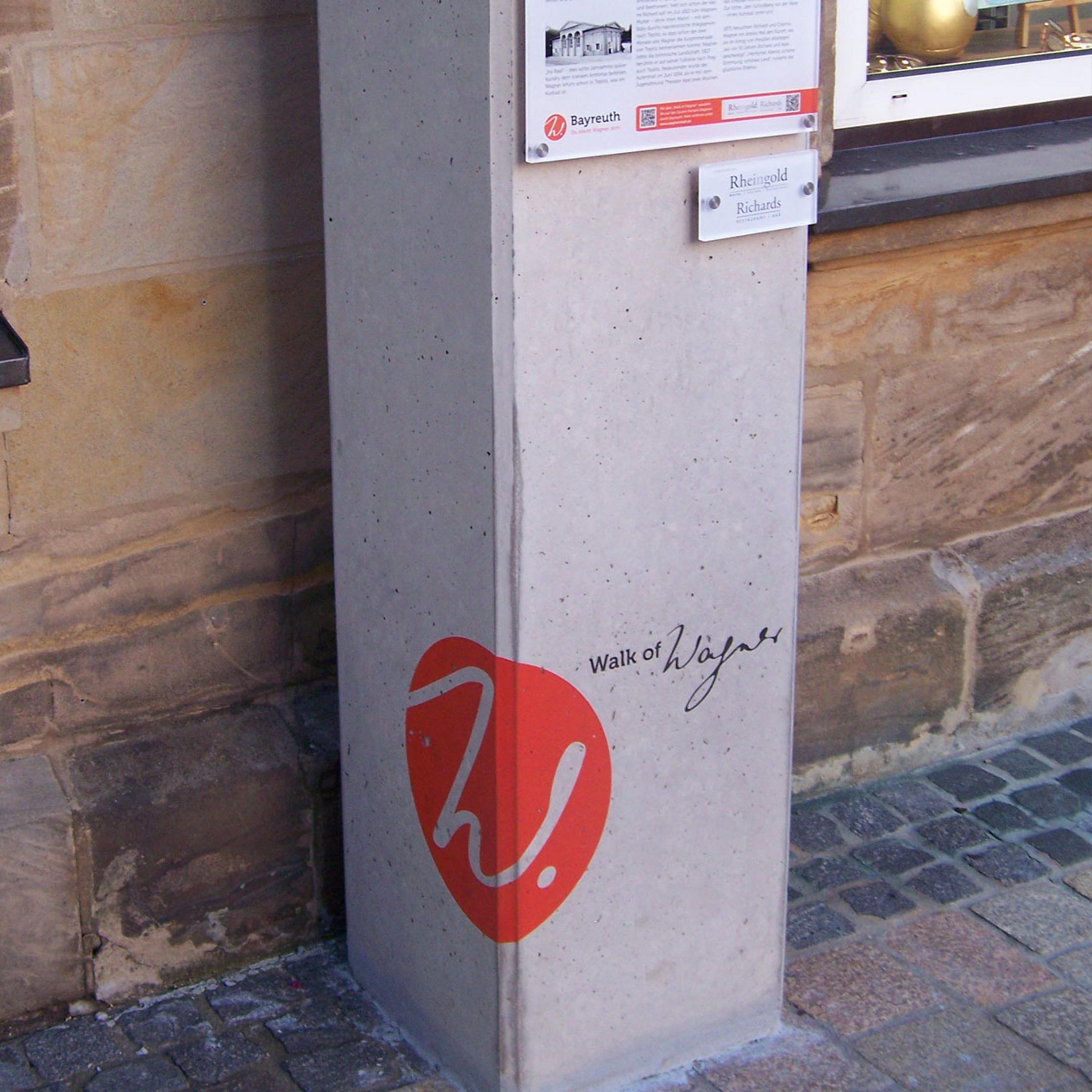 Betonsockel des „Walk of Wagner“ in Bayreuth mit Infotafel und rotem Logo an historischer Hauswand.