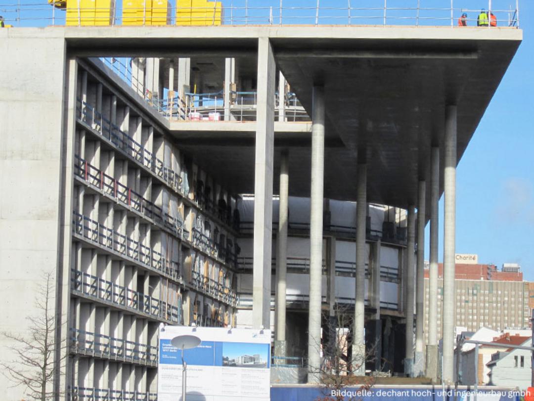 Baustelle eines mehrstöckigen Gebäude im Rohbau mit Betonpfeilern und Gerüsten, im Hintergrund ist das Gebäude der Charité zu sehen