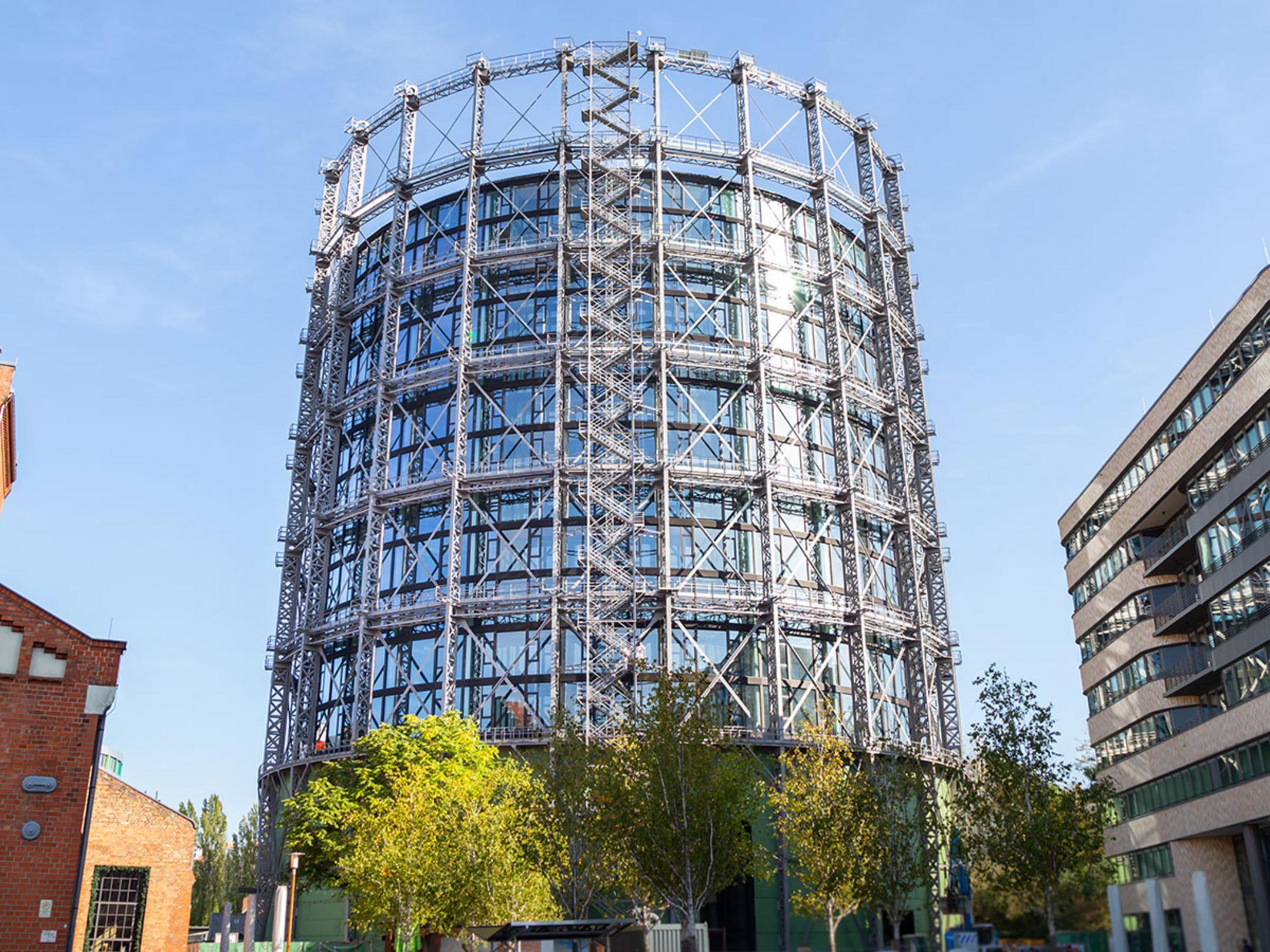 Das Gasometer vor blauem Himmel, im Vordergrund Bäume und Gebäude.