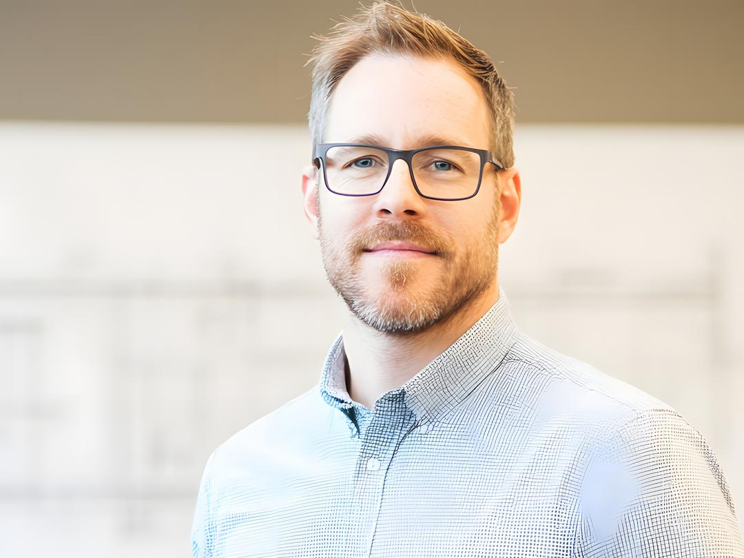 Im Vordergrund ein Mann mit Brille und Hemd, im Hintergrund unscharfs steht vor eine Tafel, auf der ein Architekturplan hängt.