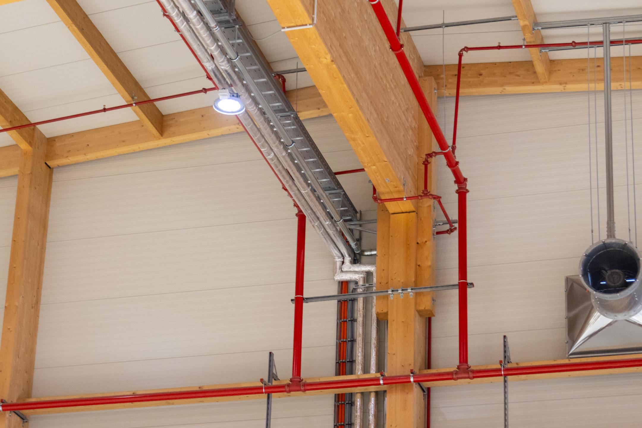 Weitspannkabelrinnen und Kabelleitern an der Decke und Wand einer Werkhalle, gepaart mit Holzbalken und roten Rohren.
