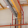 Weitspannkabelrinnen und Kabelleitern an der Decke und Wand einer Werkhalle, gepaart mit Holzbalken und roten Rohren.