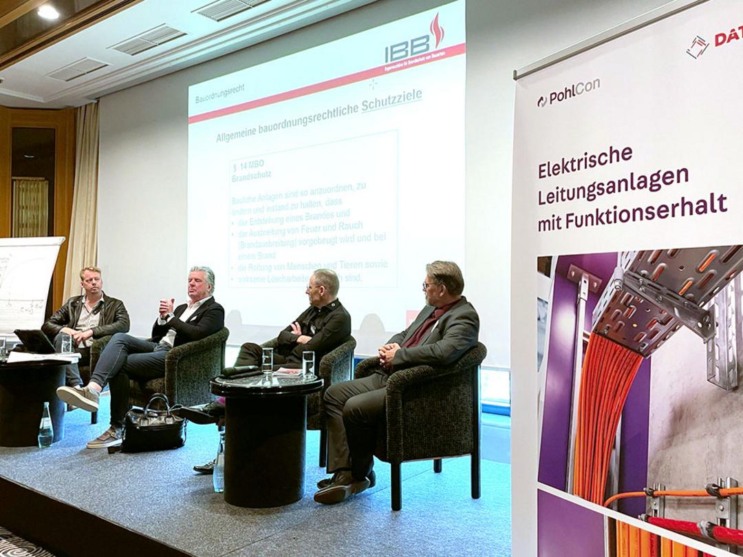 Vier Personen sitzen bei den Brandschutztagen auf einer Bühne, im Hintergrund wird eine Präsentation an die Wand projiziert.