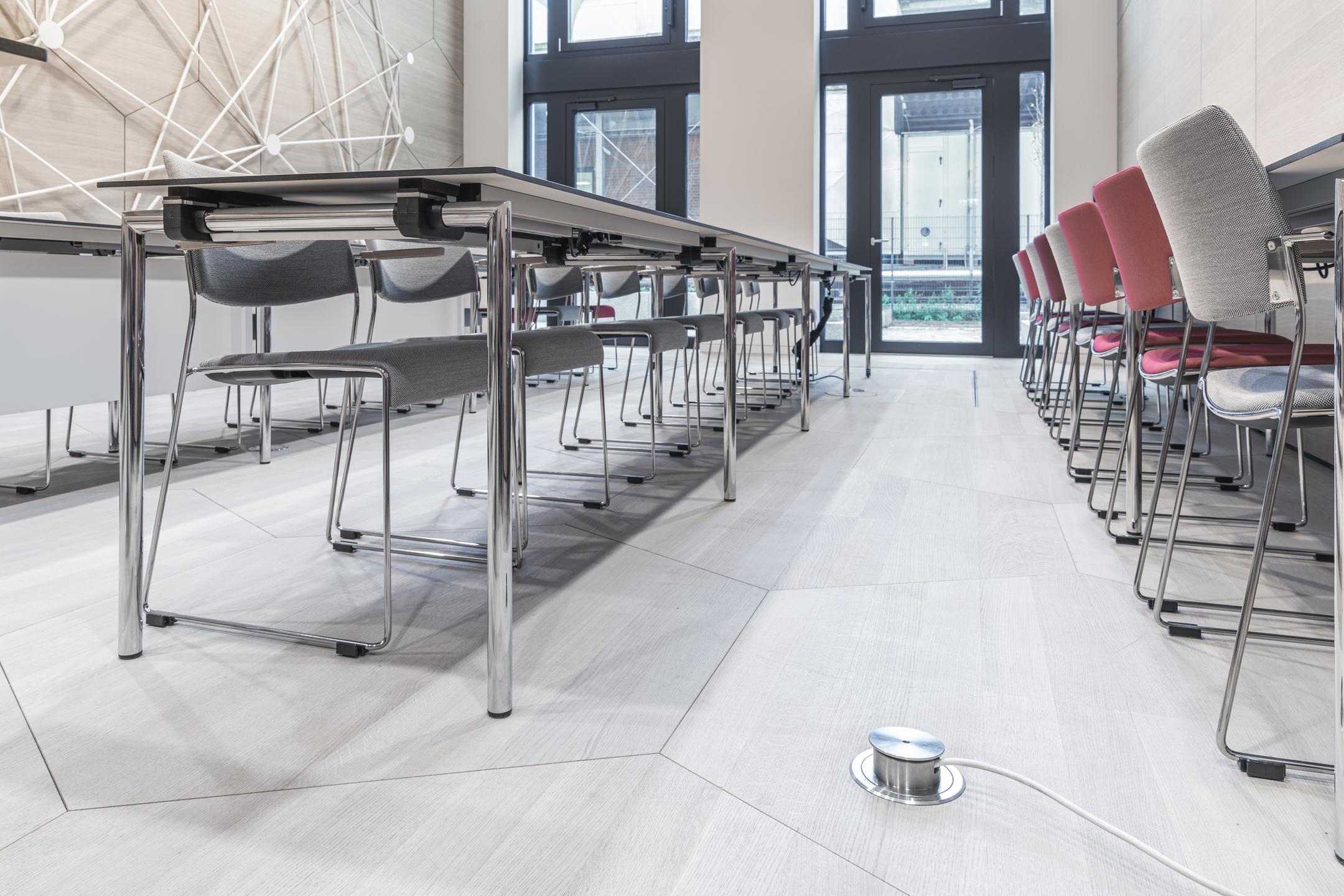 Ein moderner Arbeitsraum mit zwei langen Tischen, an denen Stühle stehen, und einem unauffälligen metallenen Kabelelement am Boden.