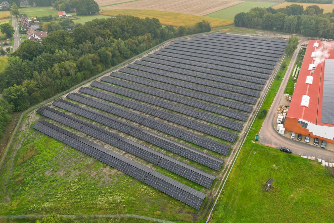 Luftaufnahme einer Photovoltaikanlage mit Feldern und Bäumen im Hintergrund, ein Industriegebäude schließt sich an.