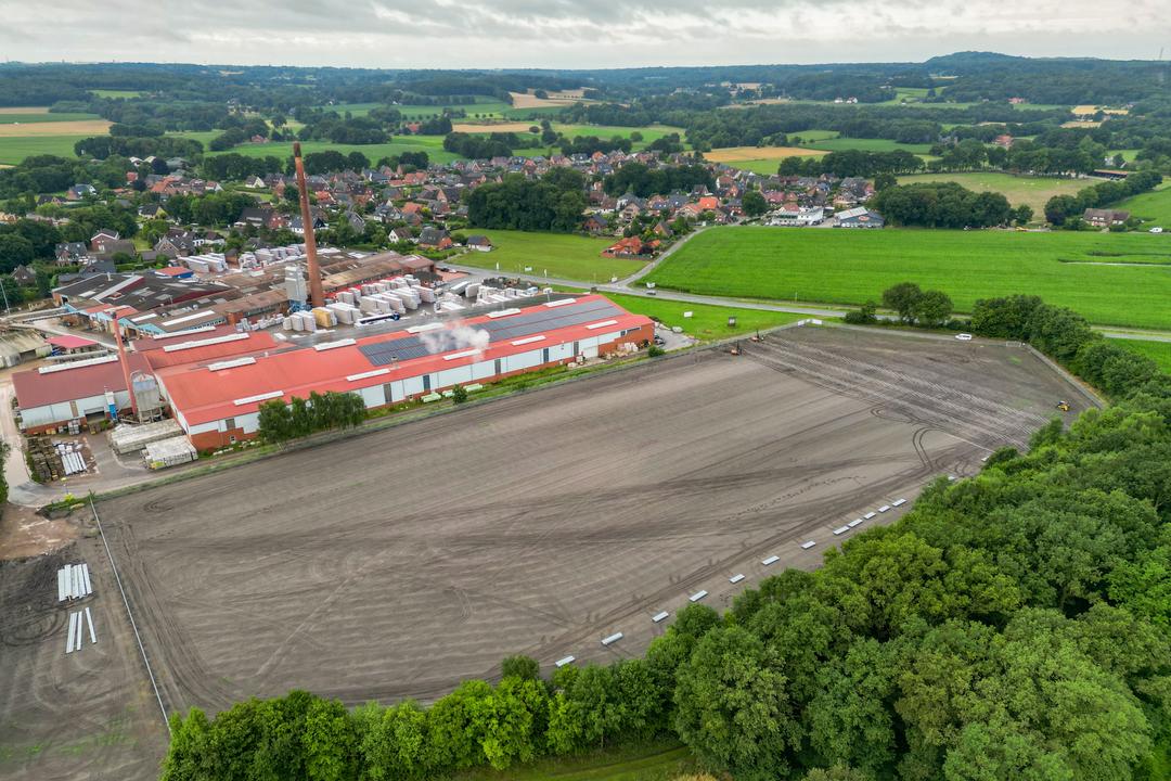 Luftaufnahme eines Industrieareals mit angrenzendem Baugelände und grüner Landschaft im Hintergrund.