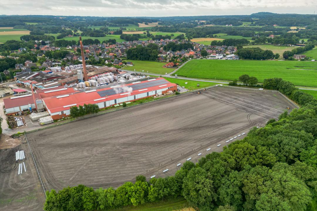 Luftaufnahme eines Industrieareals mit angrenzendem Baugelände und grüner Landschaft im Hintergrund.