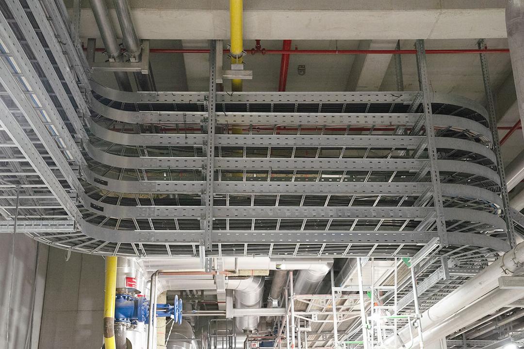 Mehrere Kabelleitern übereinander montiert und farbige Rohrleitungen verlaufen geordnet an der Decke eines Industriegebäudes