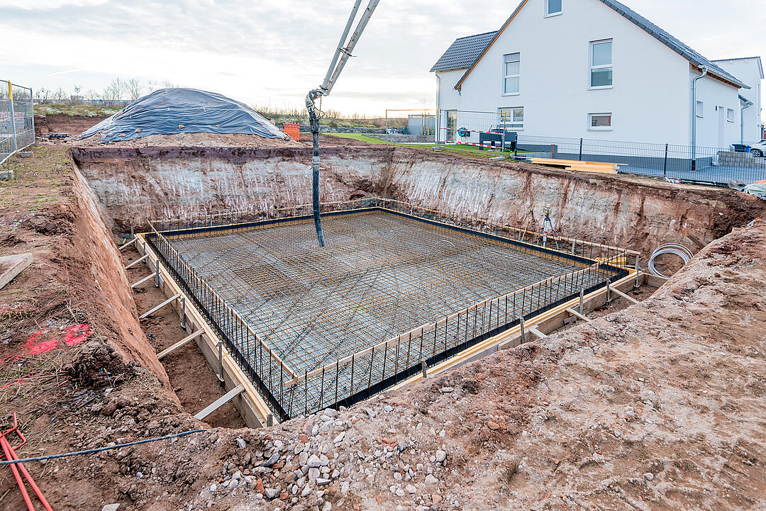 Baugrube mit bewehrter Bodenplatte vor dem Betonieren, umgeben von Schalung und aufgehender Anschlussbewehrung.