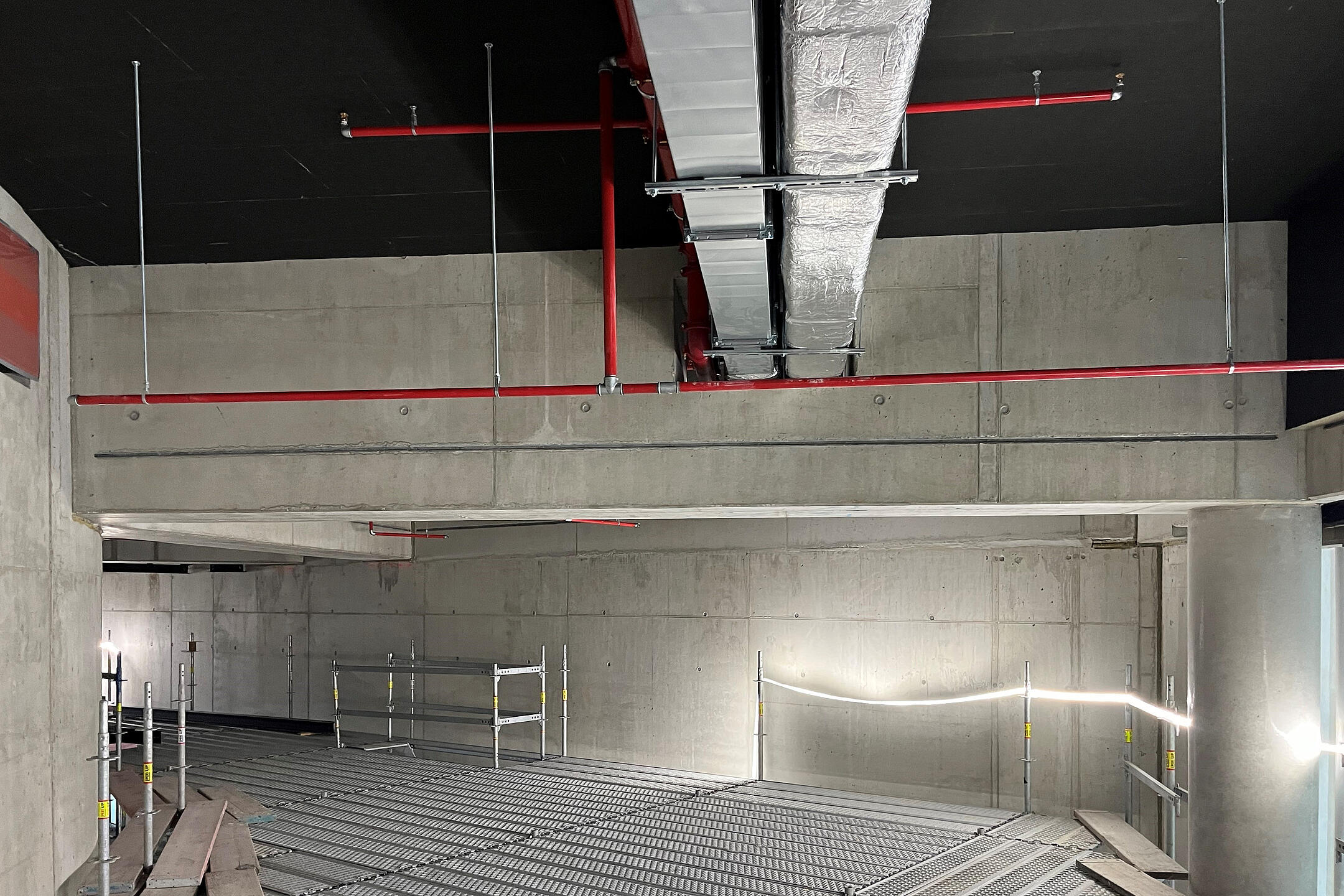 Rohbau mit abgehängten Lüftungskanälen und Sprinklerleitungen unter Betondecke, Gitterrosten als Arbeitsplattform und Gerüst.