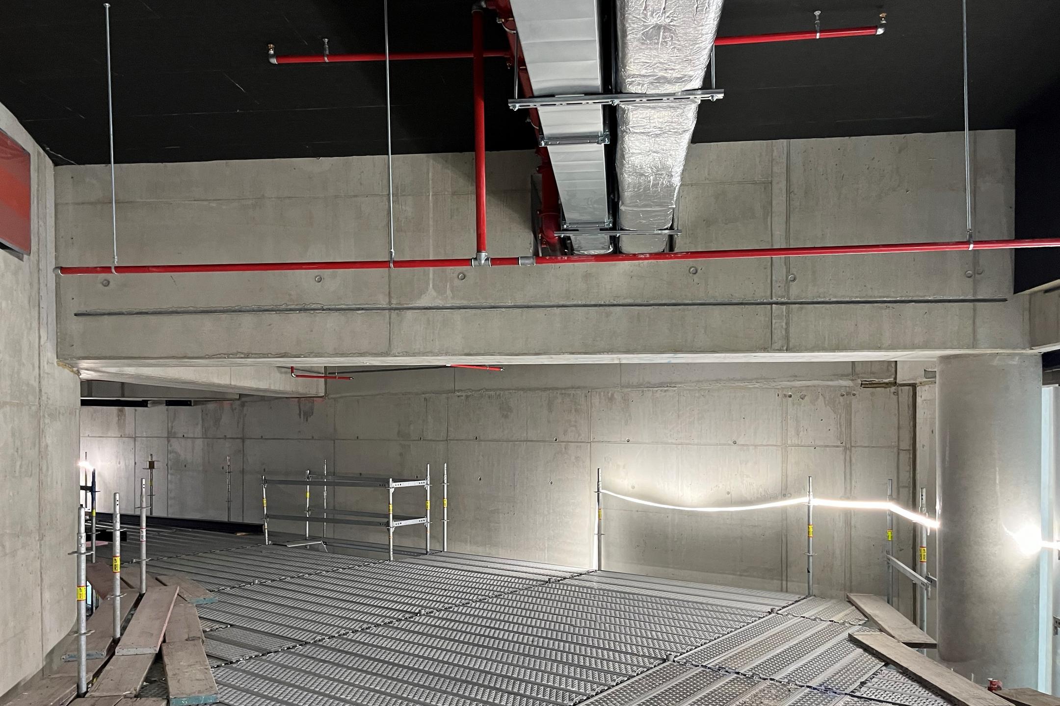 Rohbau mit abgehängten Lüftungskanälen und Sprinklerleitungen unter Betondecke, Gitterrosten als Arbeitsplattform und Gerüst.