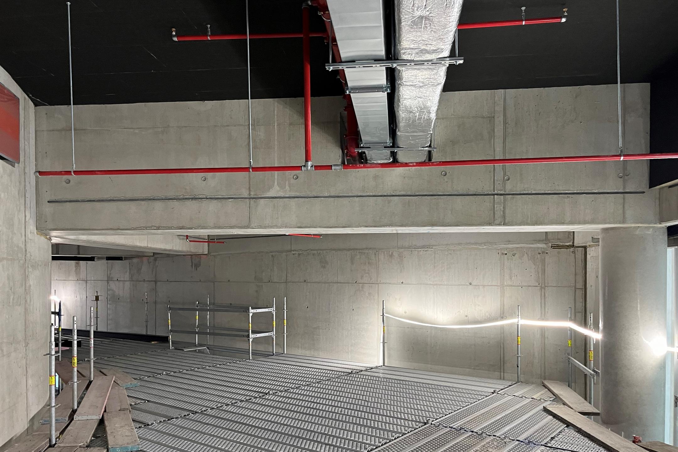 Rohbau mit abgehängten Lüftungskanälen und Sprinklerleitungen unter Betondecke, Gitterrosten als Arbeitsplattform und Gerüst.