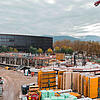 Baustelle mit zwei roten Kränen und Baumaterialen, im Hintergrund ist ein fertiges Gebäude mit Fensterfront zu sehen, bewölkter Himmel.