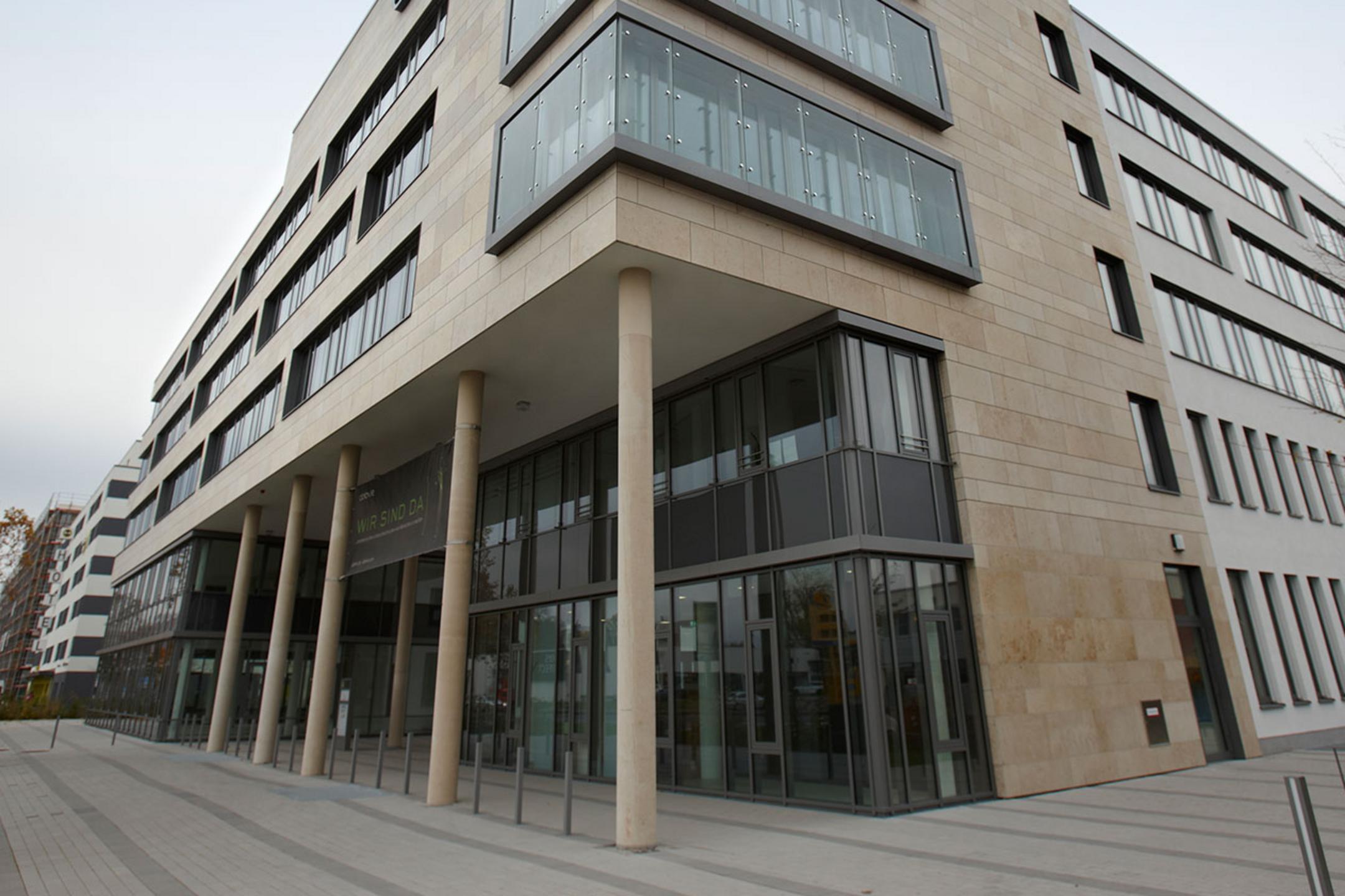Blick auf ein säulengestütztes, teilverglastes Bürogebäude, dahinter blauer Himmel.