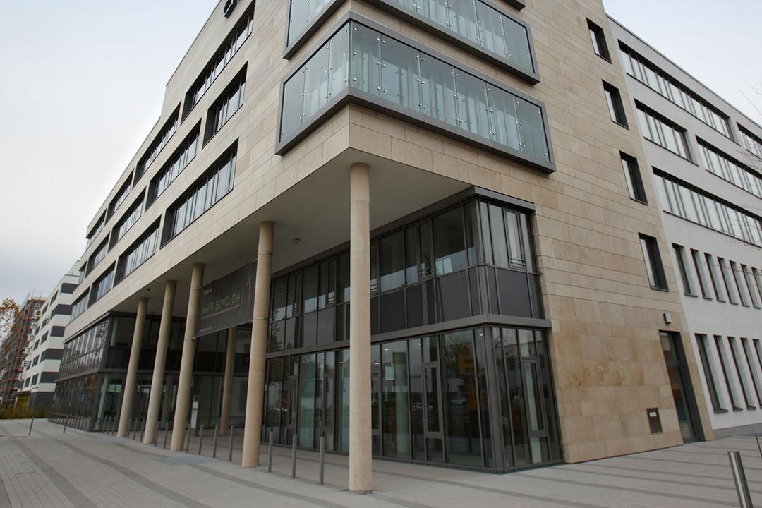 Blick auf ein säulengestütztes, teilverglastes Bürogebäude, dahinter blauer Himmel.