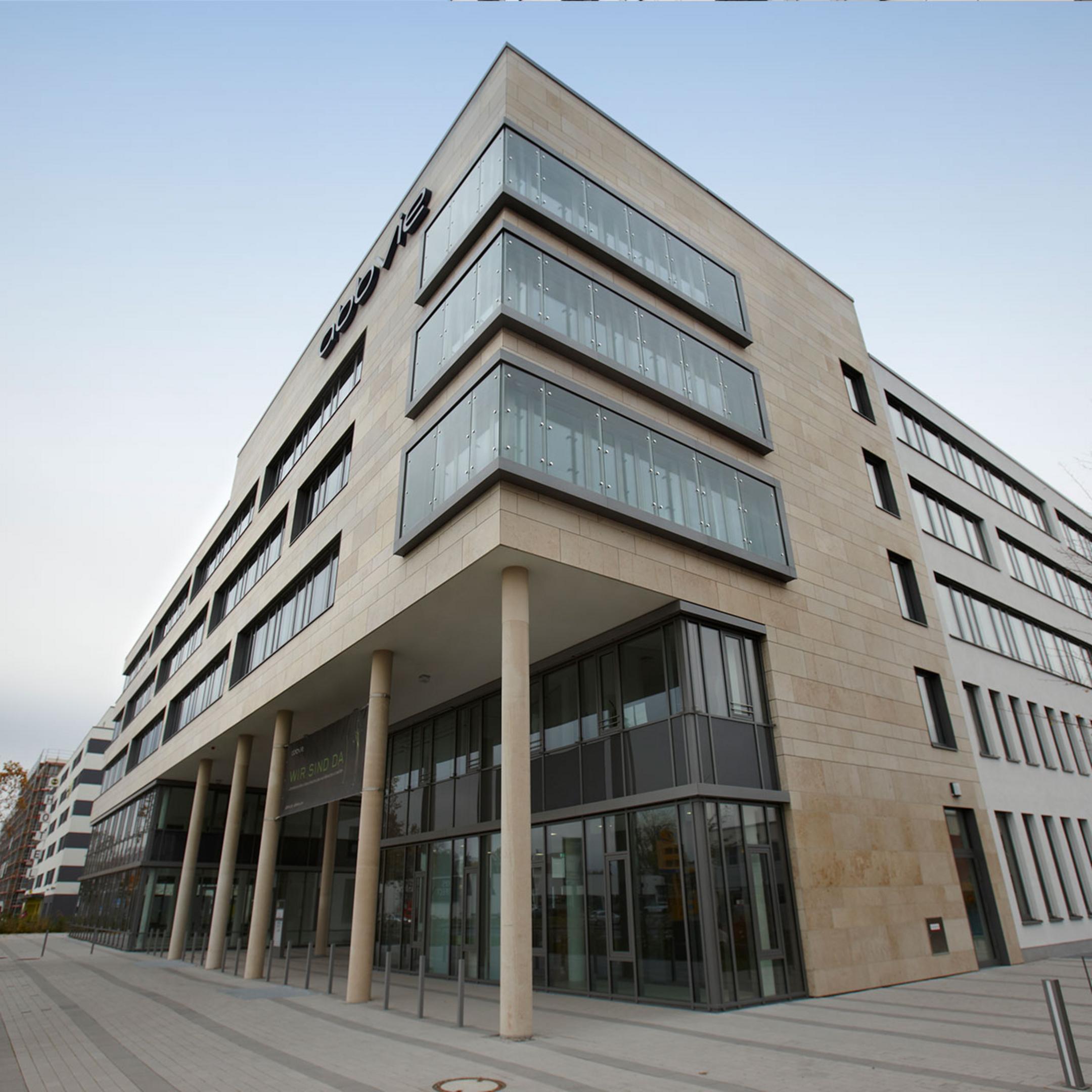 Blick auf ein säulengestütztes, teilverglastes Bürogebäude, dahinter blauer Himmel.