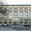 Frontalansicht Gebäude mit Fensterfront und parkenden Autos davor, ein Baum ragt auf der linken Seite ins Bild.