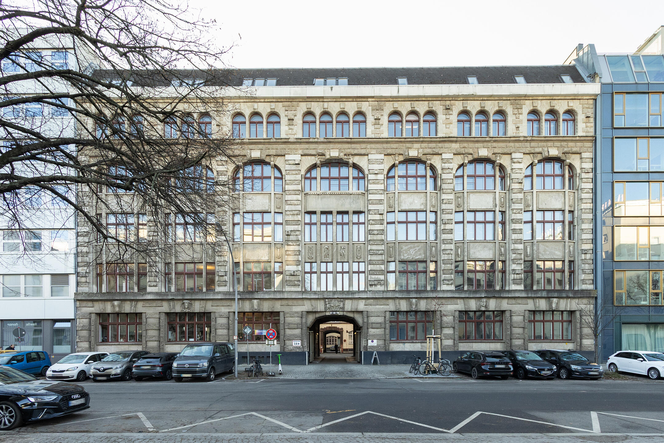 Frontalansicht Gebäude mit Fensterfront und parkenden Autos davor, ein Baum ragt auf der linken Seite ins Bild.