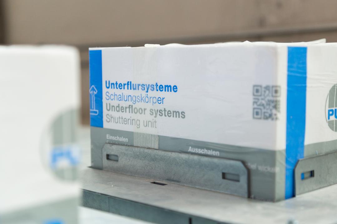 Detailansicht eines Schalungskörpers für Unterflursysteme auf einer Baustelle, beschriftet mit deutsch-englischen Titeln