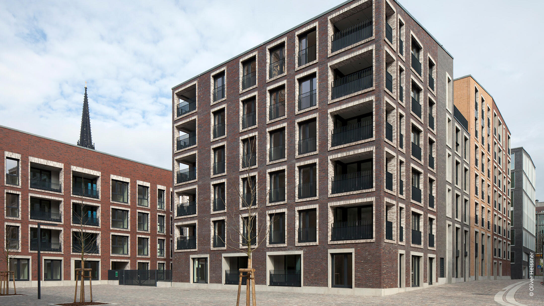 Mehrstöckiger Wohnkomplex mit Backsteinfassade, Balkonen, jungem Baumbestand und gepflastertem Vorhof