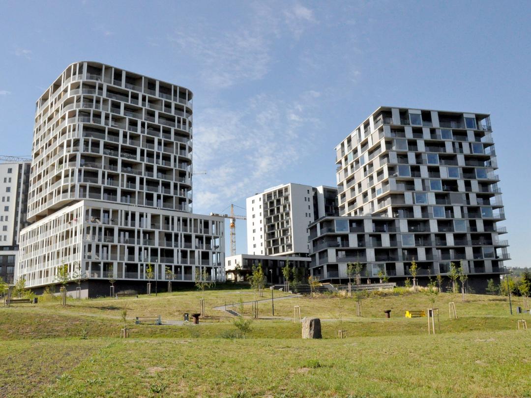 Der moderne, mosaikartige Doppel-Gebäudekomplex vor blauem Himmel, im Vordergrund Grünflächen und Bäume, aus weiterer Entfernung.