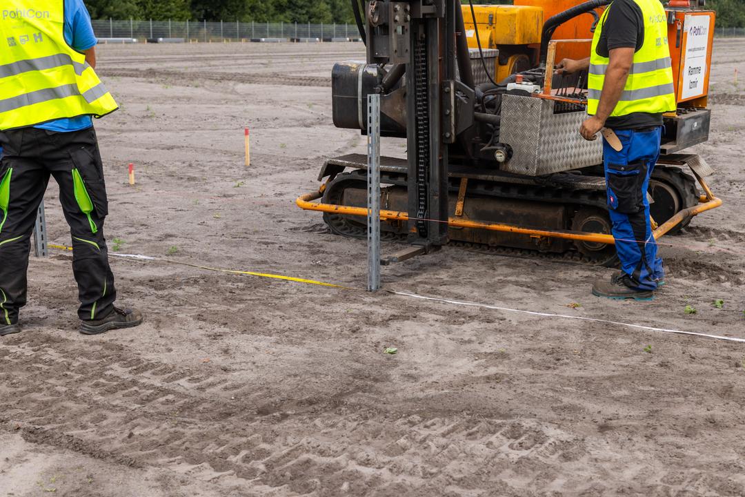 Nahaufnahme einer Baustelle mit Bauarbeitern und einem Baufahrzeig. Auf freiem Gelände werden Fundamente errichtet.