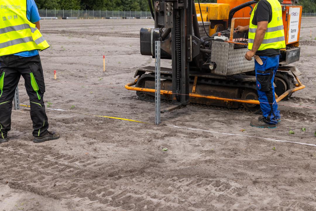 Nahaufnahme einer Baustelle mit Bauarbeitern und einem Baufahrzeig. Auf freiem Gelände werden Fundamente errichtet.