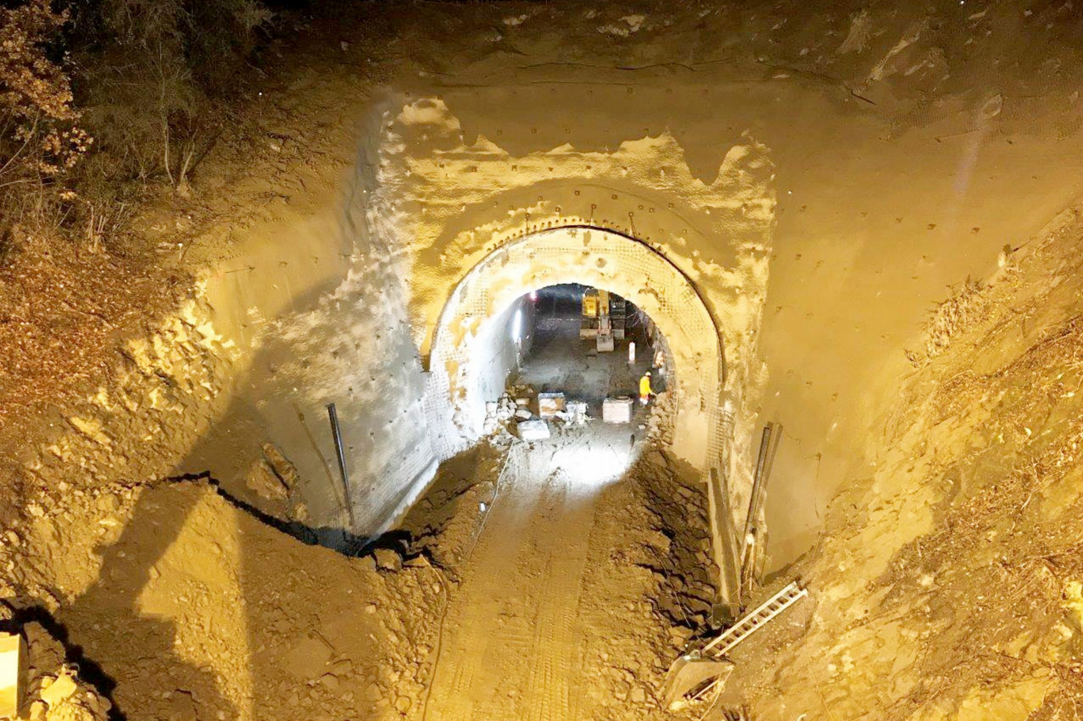 Ausgegrabener Tunnel mit beleuchtetem Eingang, Erde und Geröll, Bauarbeiter in gelber Schutzkleidung